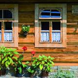 Drewniana ściana domu z podmurówką z kamieni, zbudowana z beli, z drewnianymi dwoma oknami, z wiszącymi zazdrostkami. Pomiędzy oknami i pod nimi kwiaty w doniczkach stojących na schodach z kamieni. Po prawej stronie przy oknie tabliczka z napisem 